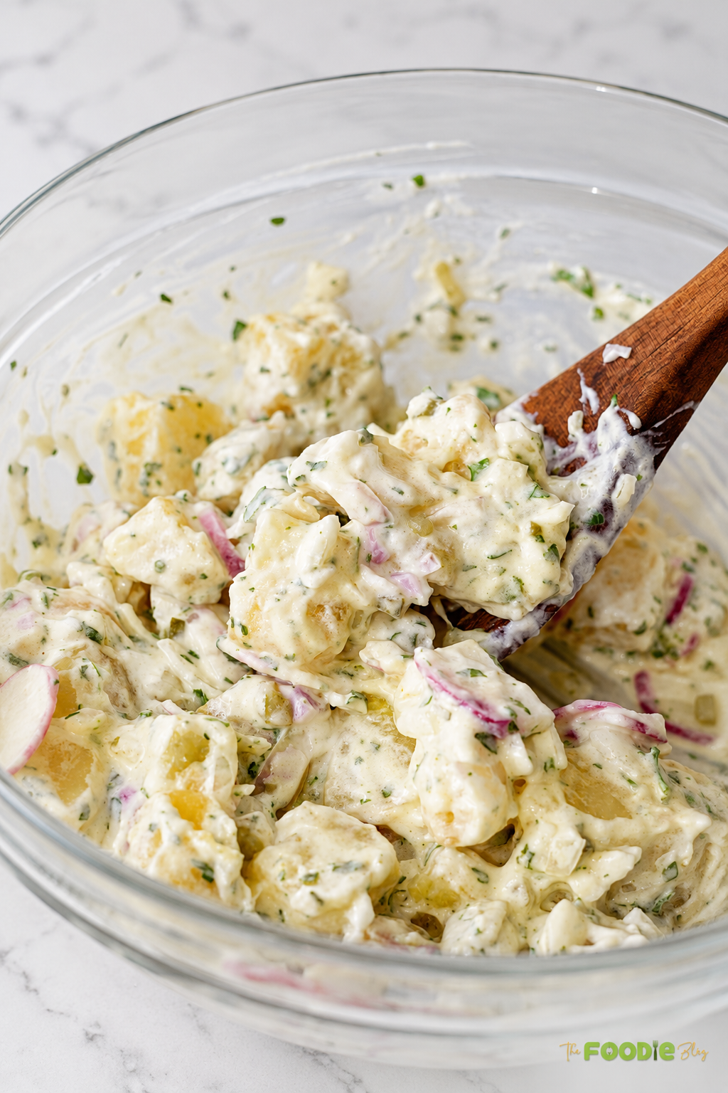 Creamy red potato salad with radishes and red onion lifted with a wooden spoon from a mixing bowl.
