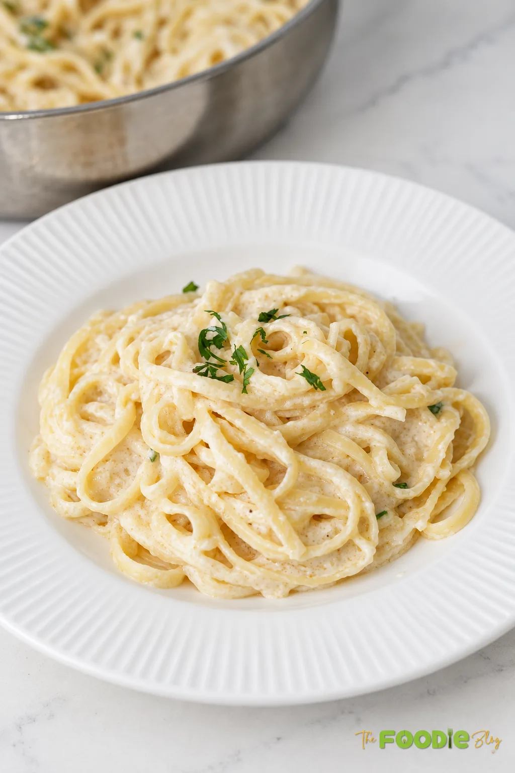 Creamy fettuccine Alfredo served in a white bowl with parsley and a skillet of pasta in the background.
