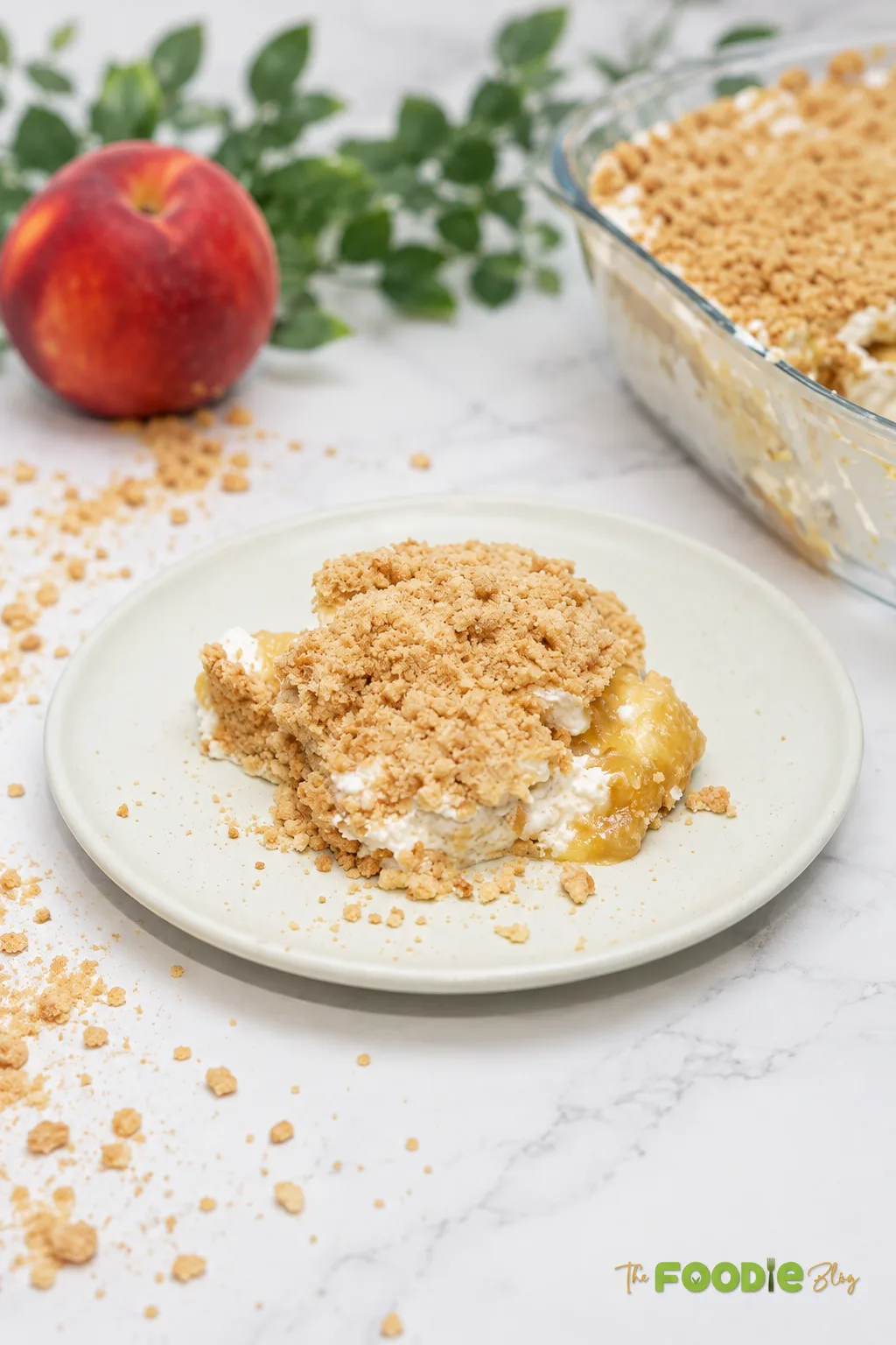 Serving of creamy no-bake peach dessert on a plate with biscuit crumbs and fresh peach in the background.